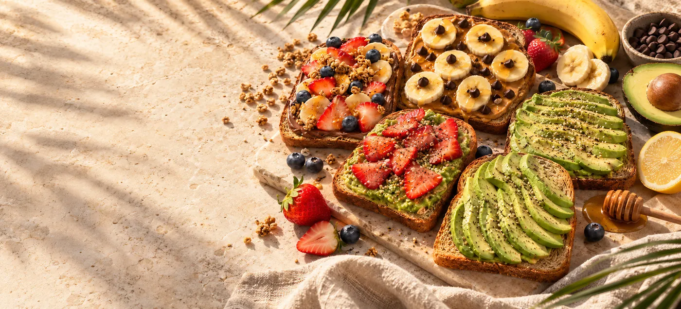 Toasts with avocado, strawberries, banana and chocolate chips overlapping on pale stone in warm side sun, with a halved avocado, fresh fruit and a honey dipper